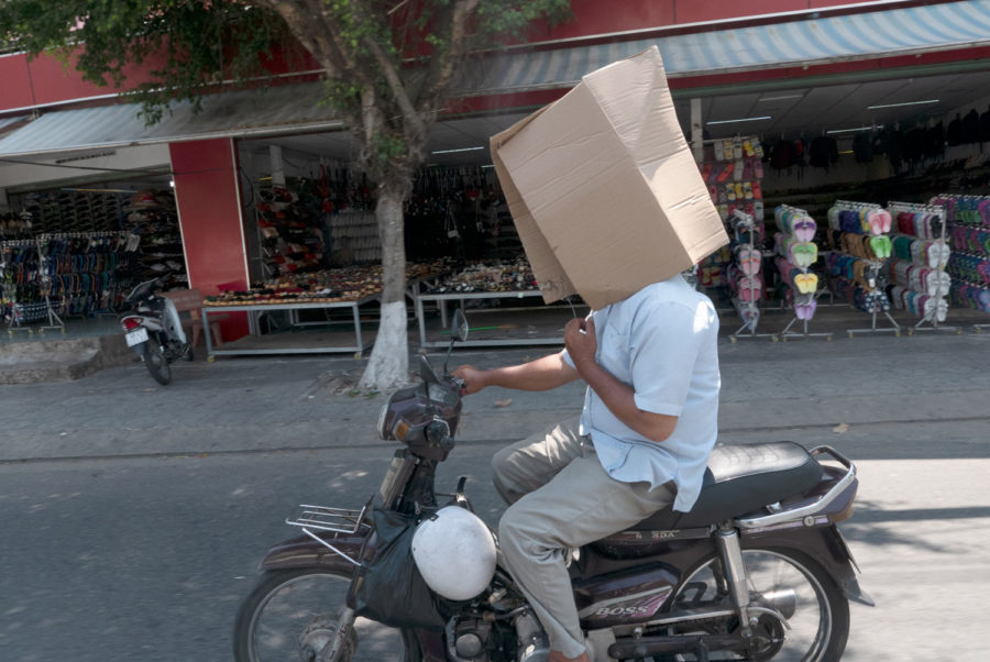Un hombre conduce una moto en Asia con una caja en la cabeza para protegerse del sol. Motos en Asia