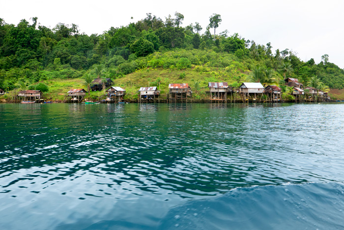 Unas 11 casa se levantan de la orilla de unos 5 metros con postes de madera. Los tejados son de uralita metálica. Al fondo la selva. el terreno tiene mucha pendiente. RAJA AMPAT. PAPÚA. INDONESIA.