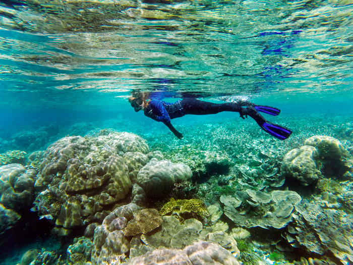 Una buceadora mira hacia cámara mientras pasa por encima de un montón de corales duros. Una persona alza los brazos en cruz en medio de una islote de arena rodeado de un mar turquesa. RAJA AMPAT. PAPÚA. INDONESIA.