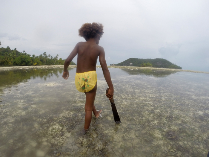 Una niña aborigen con un machete camina por el mar en busca de conchas y tiburones pequeños. Una persona alza los brazos en cruz en medio de una islote de arena rodeado de un mar turquesa. RAJA AMPAT. PAPÚA. INDONESIA