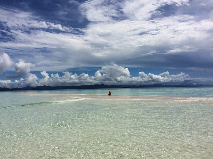En medio de un saliente de arena totalmente rodeado de mar se sienta una mujer tomando el sol. El cielo esta lleno de nubes de algodón y un cielo azul. Una persona alza los brazos en cruz en medio de una islote de arena rodeado de un mar turquesa. RAJA AMPAT. PAPÚA. INDONESIA.