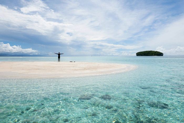 Una persona alza los brazos en cruz en medio de una islote de arena rodeado de un mar turquesa. RAJA AMPAT. PAPÚA. INDONESIA