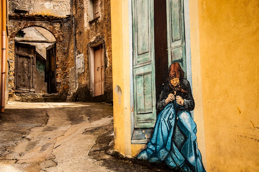 Preciosa calle donde vemos una pintural mural con una señora cosiendo en la puerta de su casa. Orgosolo. Cerdeña.