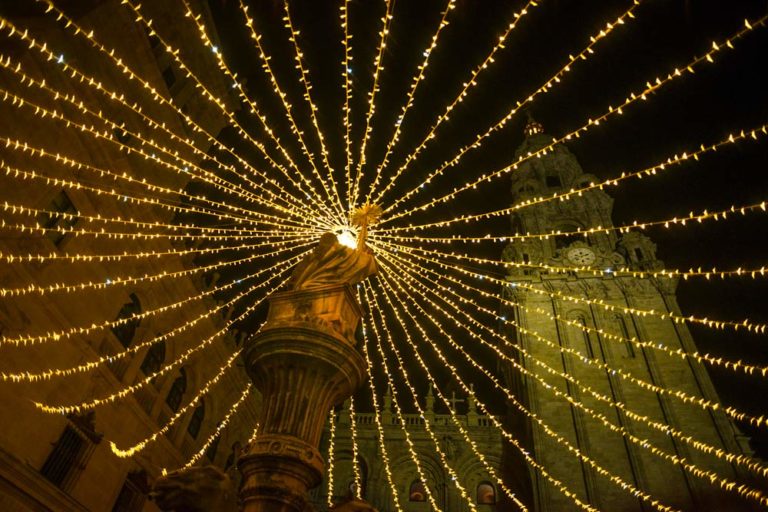 Luces a modo de tela araña encima de la estatua de una plaza, Una torre de la catedral se ve al fondo. Navidad en Galicia. 8 Costumbres y tradiciones. Perder el Rumbo