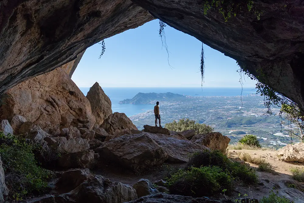 una persona sobre una roca vista desde dentro de el Forat. LA silueta de la cueva enmarca Albir y la ciudad de Benidorm.