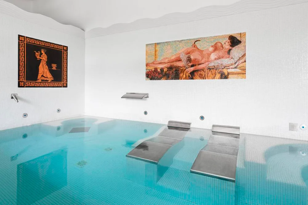 Dos tumbonas de metal en el interior de un spa. las paredes blancas están decoradas con mosaicos clásicos romanos. el agua de color turquesa.