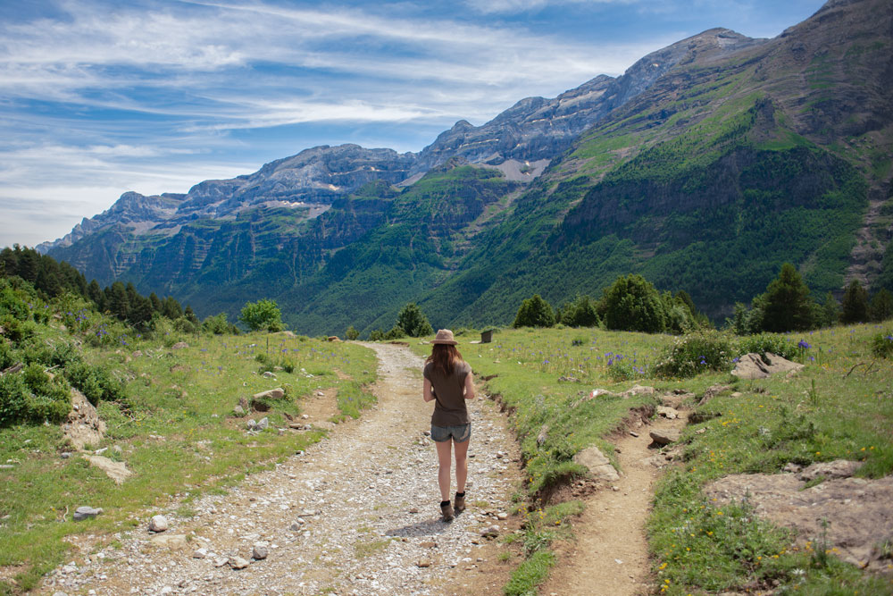 Una chica camina por un sendero en la parte alta de una cadena montañosa. Al fondo vemos impresionantes montañas llenas de verde y las partes altas de nieve