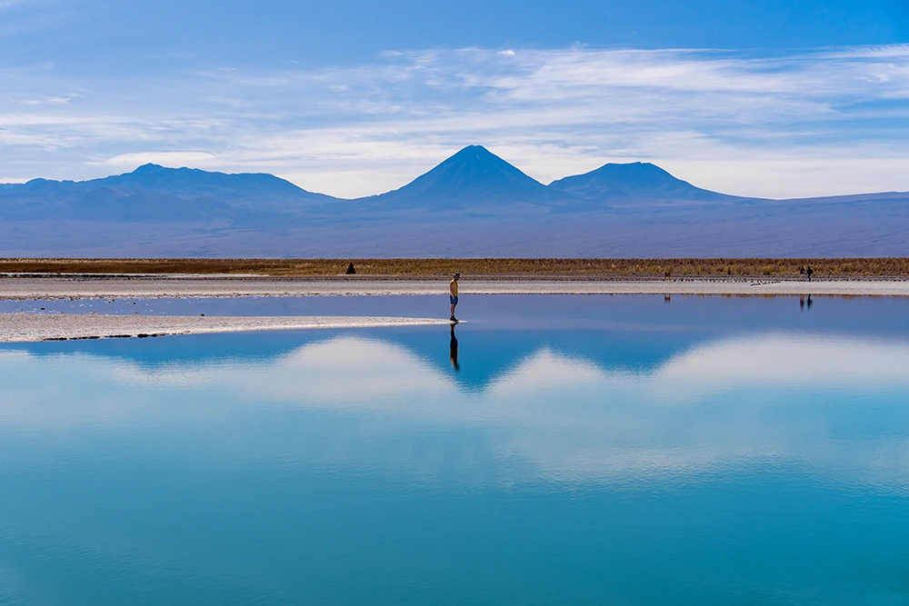Una persona se refleja en el azul turquesa de un laguna salada. Al fondo vemos volcanes y una cordillera montañosa.