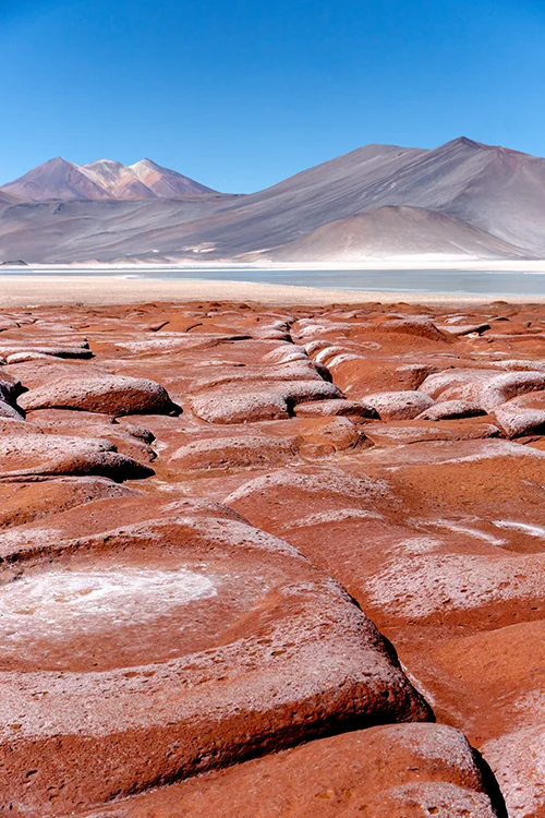 Unas piedras rojas en primer trémino dan pie a unas montañas grisáceas con una laguna turquesa en sus bases. Un bonito lugar que ver en el desierto de atacama.