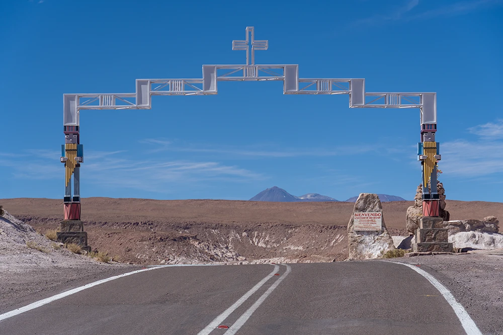 Un arco de metal cabezado por una cruz y sostenido en las cabezas de unos muñecos imitando a unos incas tocando flautas. Hay un cartel que nos da la bienvenida.