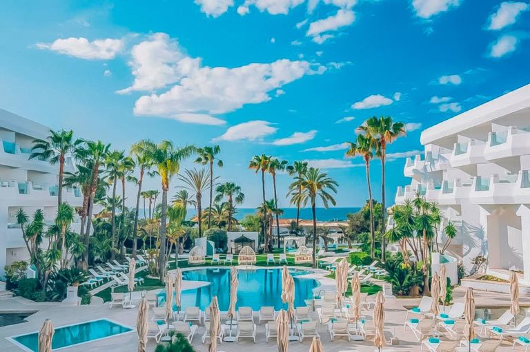 Una piscina de color azul turquesa rodeada de palmeras a ambos lados de la piscina se encuentran unas instalaciones hoteleras al fondo la playa y el mar. Iberostar Málaga. Hoteles para unas vacaciones en la playa.
