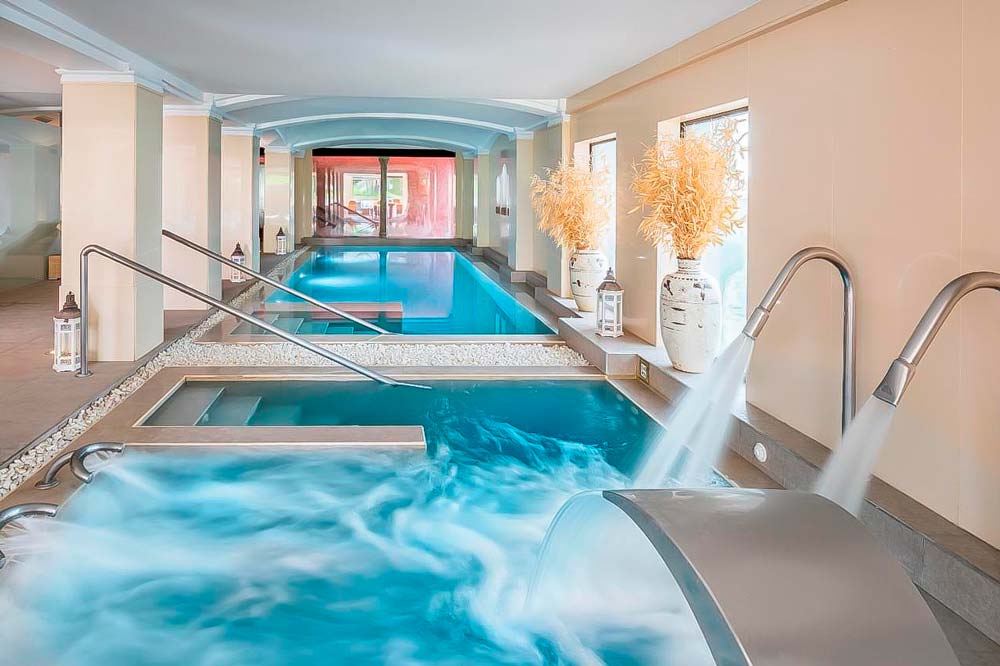 Se ve un spa de aguas cristalinas y azules con varios chorros y grandes ventanales. Hoteles con spa en España