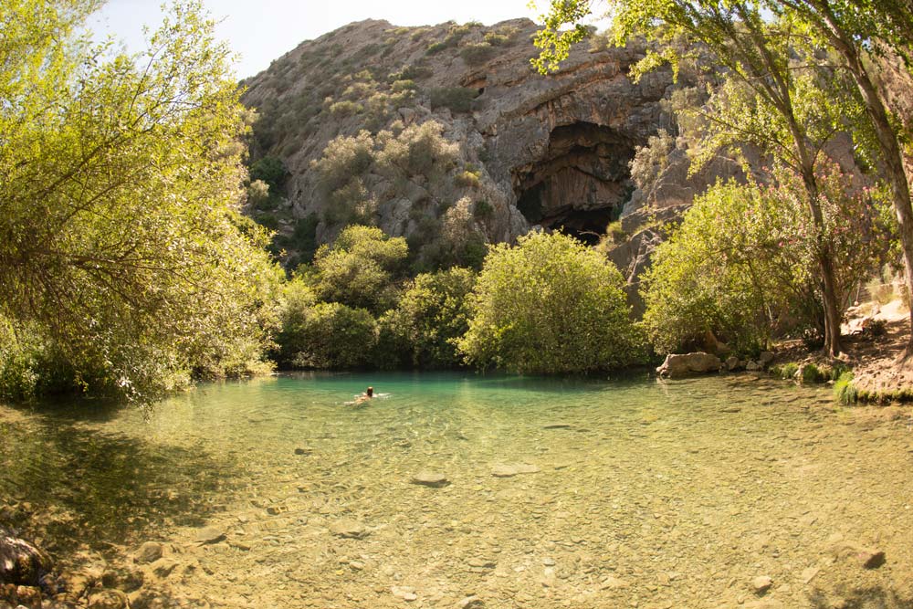Fotografía donde se ve la parte exterior de la Cueva del Gato en Málaga. Hay una enorme piscina natural de aguas cristalinas rodeada por vegetación.