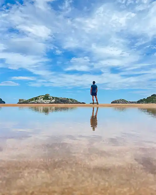Un chico observa unos islotes y su figura se refleja en un gran charco de mar.