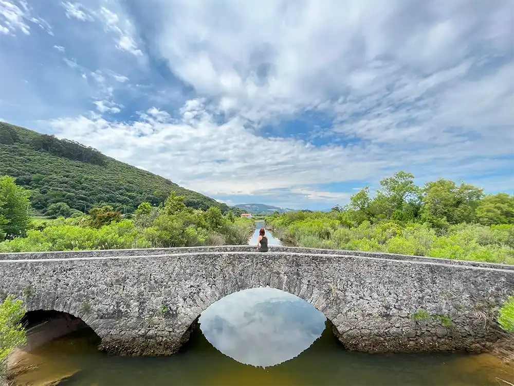Una chicas se encuentra sentada en medio de un puente romano de 2 ojos. Al fondo un lago y una montaña llena de matorrales.