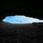 La Cueva de la Ojerada, se ve la figura de una chica en el centro de una ce las aberturas y el cielo azul al fondo