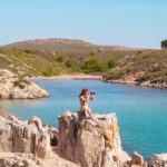 Se ve en primer término a una chica subida a una roca y haciendo una fotografía. Al fondo hay una preciosa cala de arena blanca y aguas turquesas.