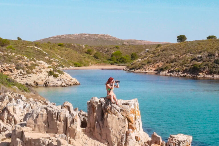 Se ve en primer término a una chica subida a una roca y haciendo una fotografía. Al fondo hay una preciosa cala de arena blanca y aguas turquesas.