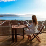 Chica que toma un café en la terraza de madera de una de las cabañas en A Costa da Morte con vistas a una preciosa playa en un día de sol y cielo azul radiante.