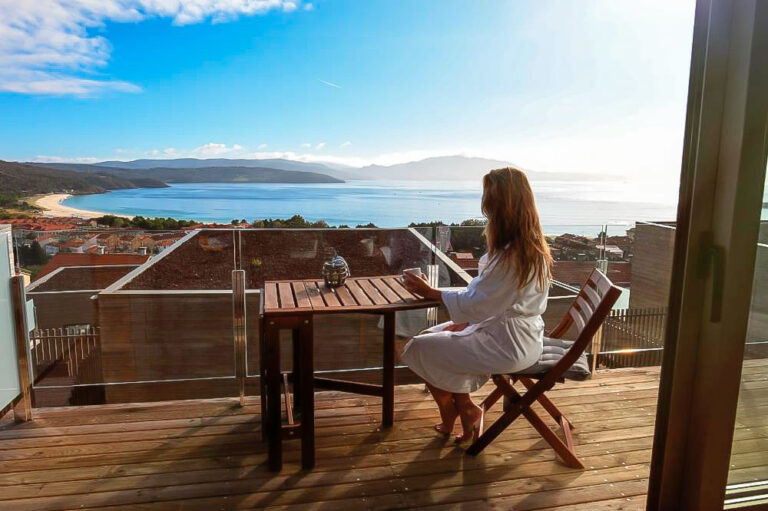 Chica que toma un café en la terraza de madera de una de las cabañas en A Costa da Morte con vistas a una preciosa playa en un día de sol y cielo azul radiante.