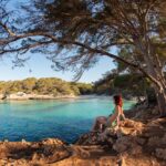 Chica sentada en unas rocas en bañador contemplando el mar azul turquesa y una playa al fondo. Lugares para viajar a Menorca maravillosos.