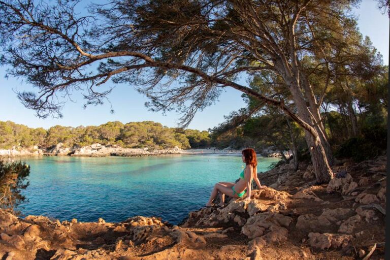 Chica sentada en unas rocas en bañador contemplando el mar azul turquesa y una playa al fondo. Lugares para viajar a Menorca maravillosos.